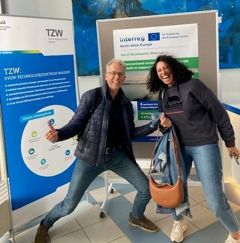 Two participants smiling and posing in front of information posters from TZW and the Interreg NWE Rural Roadwater Rescue project.
